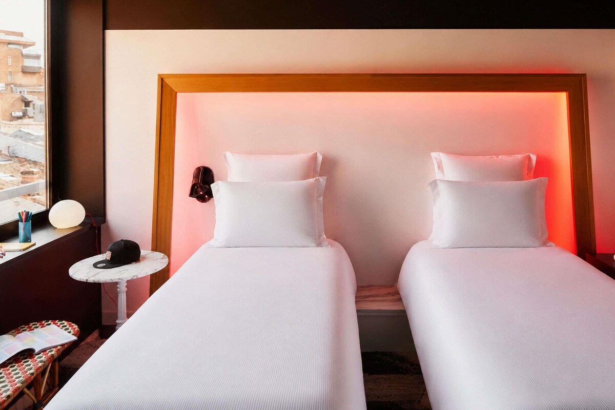 Habitación de hotel con dos camas, una mesita con lámpara, una silla y vistas a la ciudad.