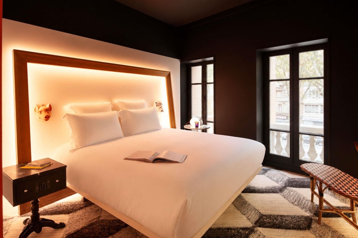 Habitación de hotel con una cama grande, una mesita de noche con lámpara, una silla y una ventana con vistas a edificios.