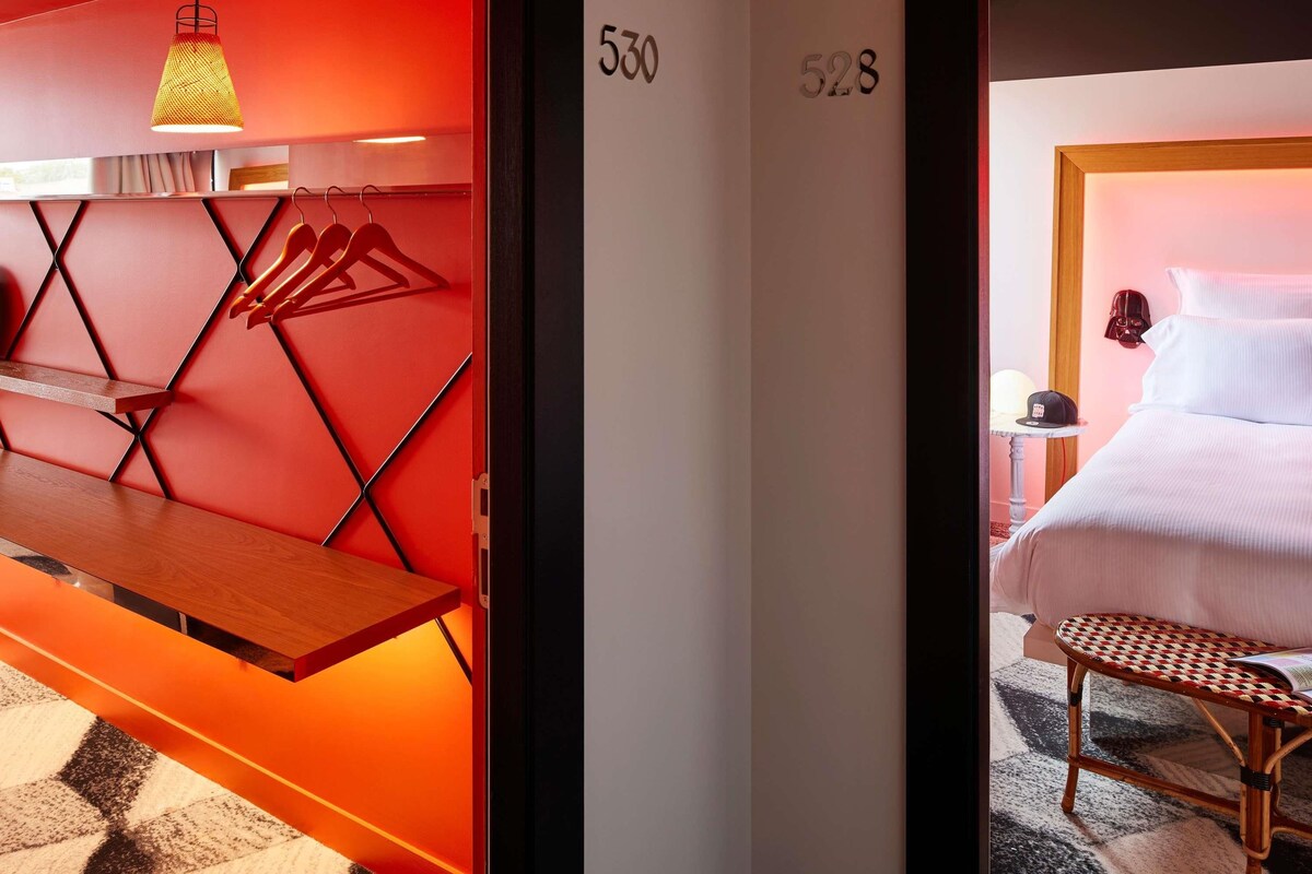 Una habitación de hotel con una pared de acento roja, una cama con ropa blanca, un banco de madera, un perchero en la pared y una lámpara colgante.