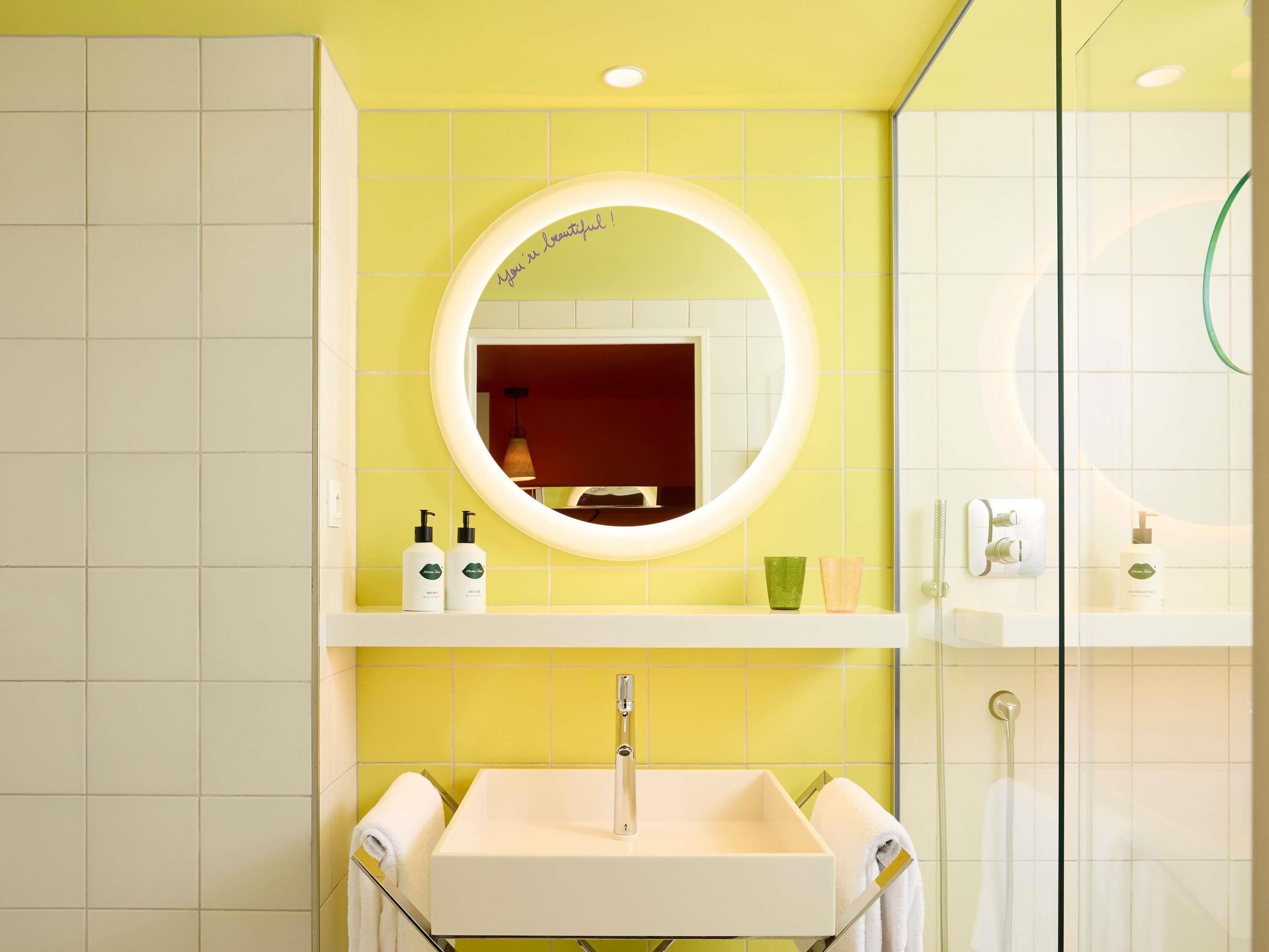 A bathroom with a yellow tiled wall, a round mirror, a white sink, and a glass shower enclosure.