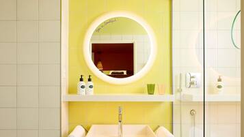 A bathroom with a yellow tiled wall, a round mirror, a white sink, and a glass shower enclosure.
