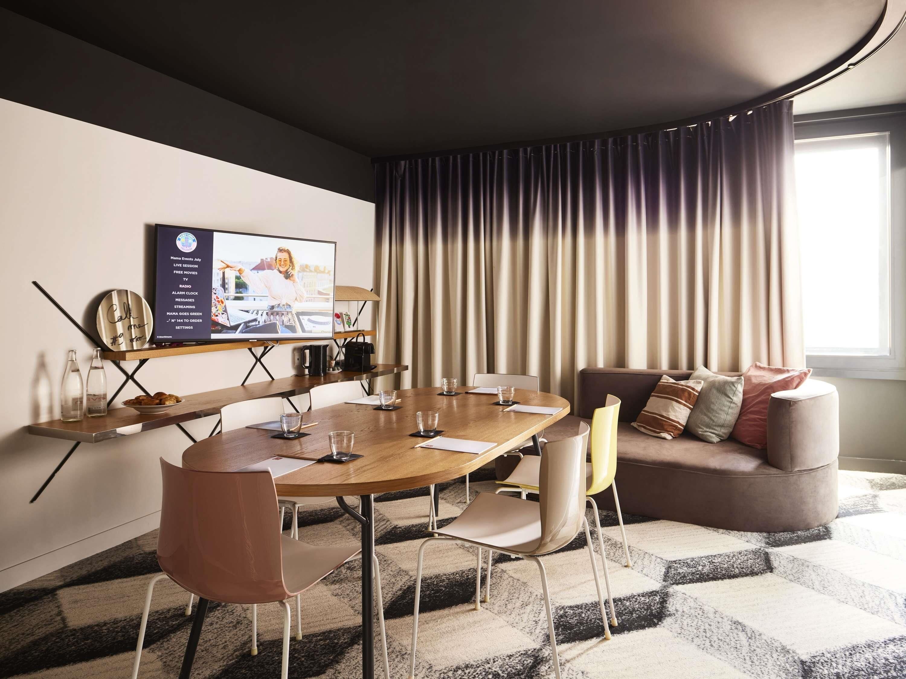 Une chambre d’hôtel moderne avec une table de salle à manger ronde, des chaises, un canapé et un téléviseur diffusant un programme en direct.