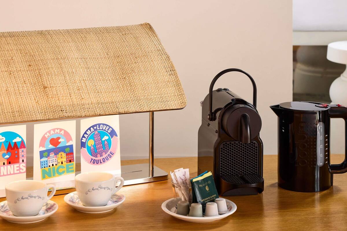 Una mesa con una lámpara, un altavoz portátil, una cafetera y un plato con cubitos de azúcar y una tarjeta.