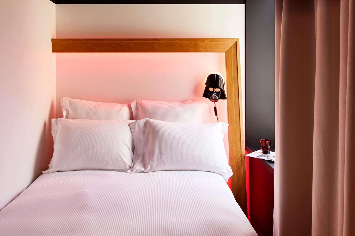 Una habitación de hotel con una cama, una mesita de noche, una lámpara y una cortina.