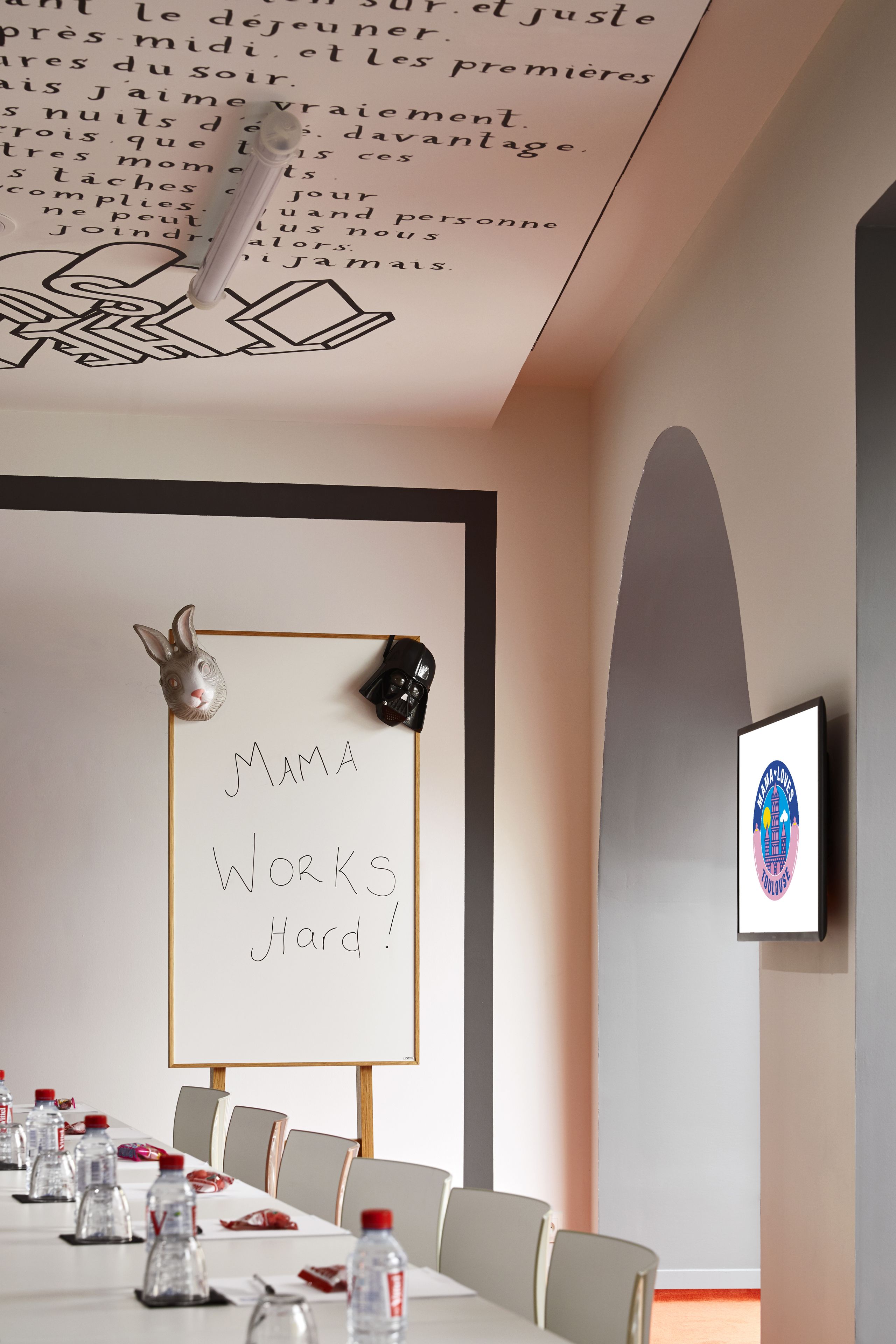 Una sala de conferencias con una pizarra mostrando 'MAMA WORKS Hard!' y un logotipo enmarcado en la pared.