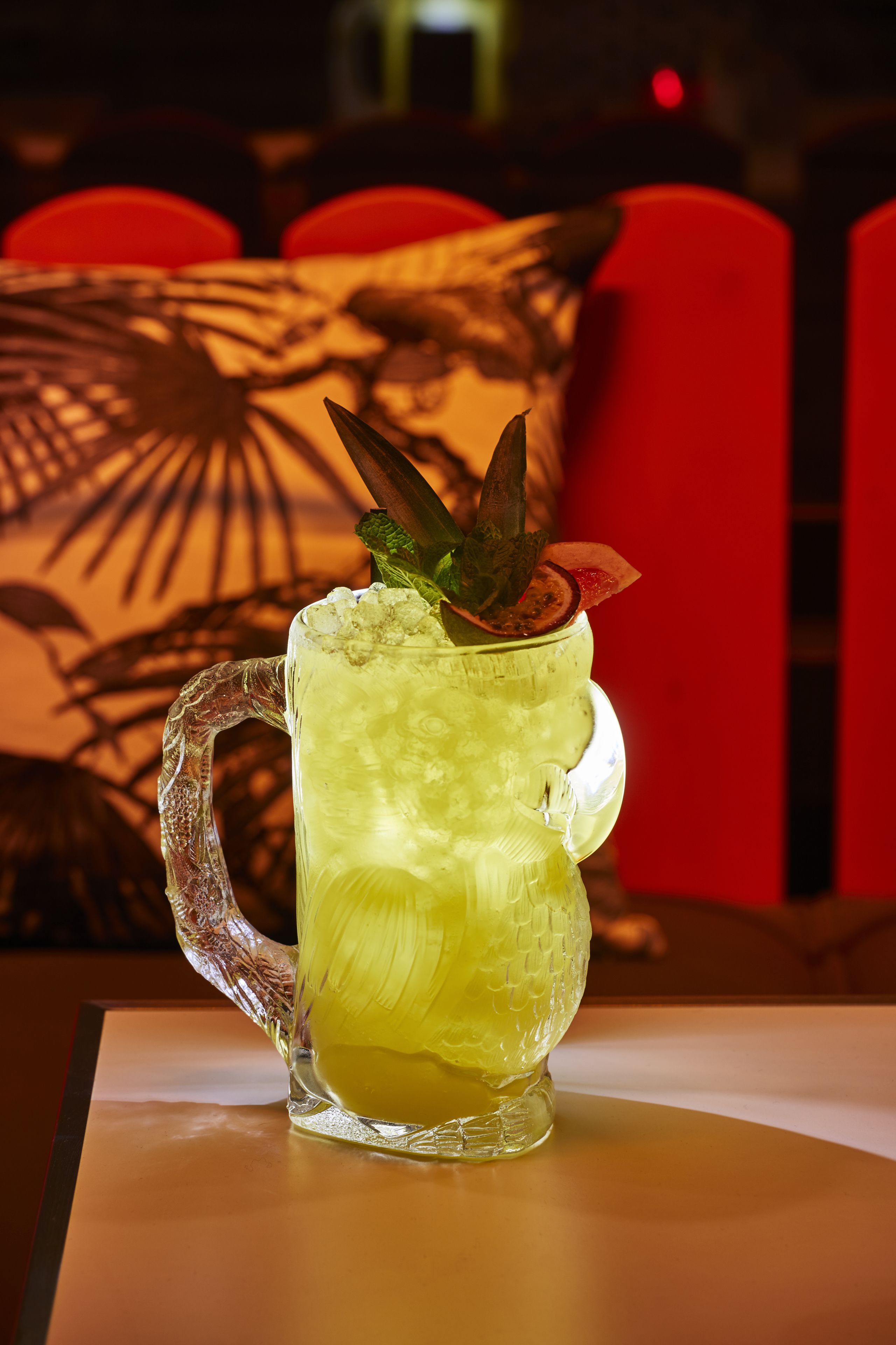 A clear glass mug with a handle, filled with a yellow beverage, garnished with a mint leaf and a slice of fruit, placed on a table with a red chair in the background.