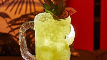 A clear glass mug with a handle, filled with a yellow beverage, garnished with a mint leaf and a slice of fruit, placed on a table with a red chair in the background.