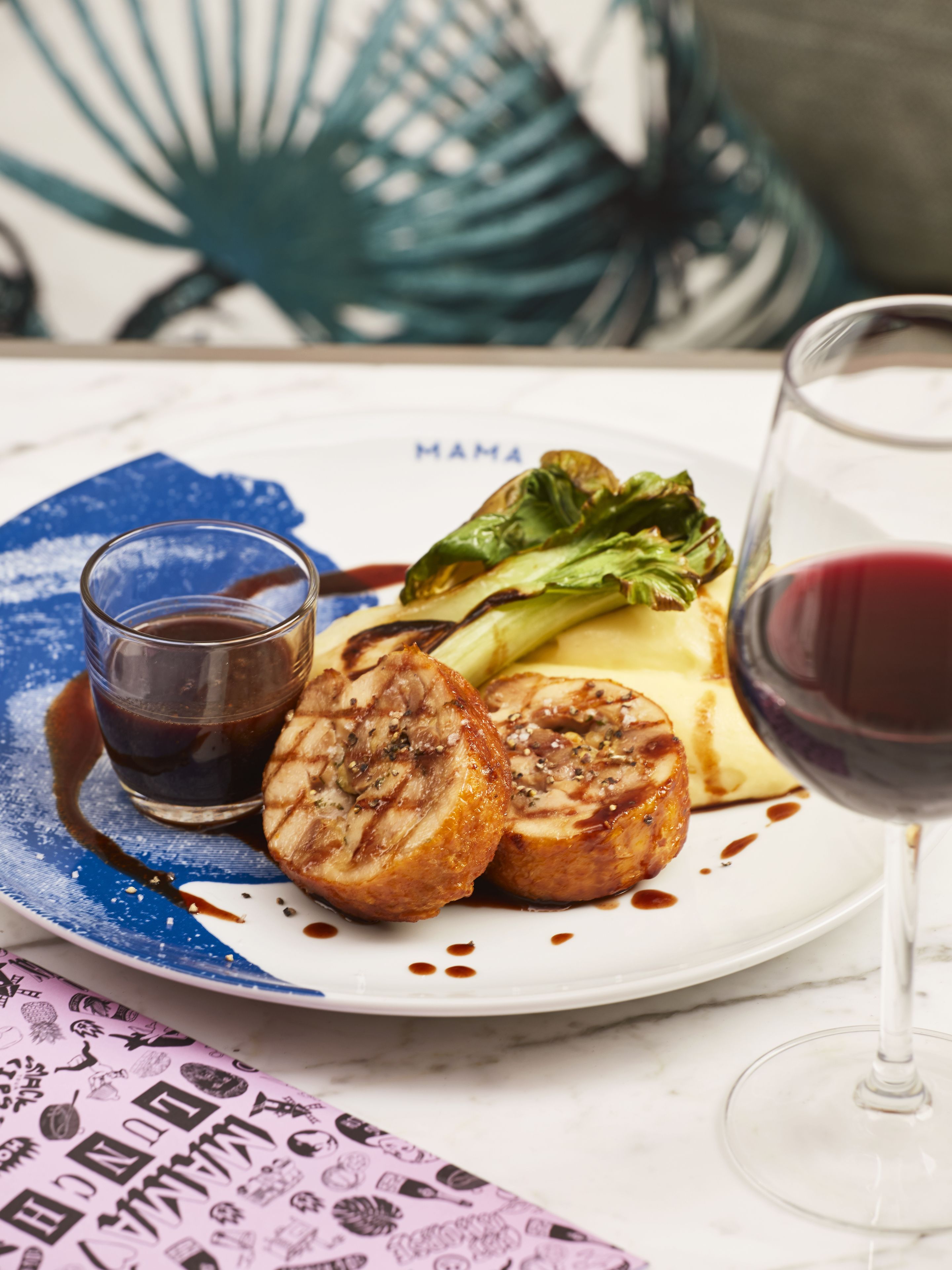 A plate with grilled meat, vegetables, and a glass of red wine on a table with a blue napkin and a patterned placemat.