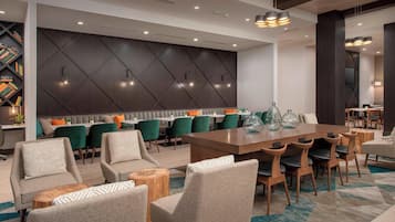 A modern hotel lobby with a long dining table, comfortable seating, and a decorative wall.