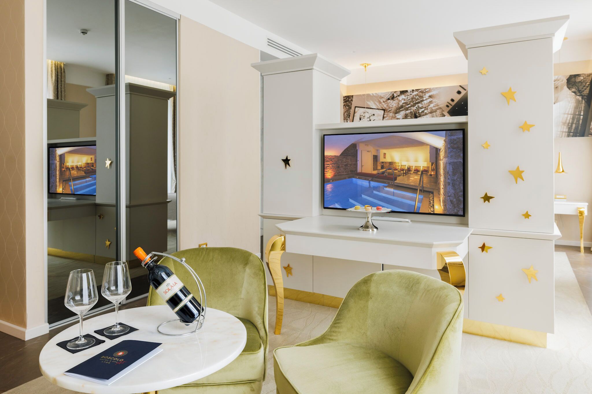 Habitación de hotel con televisión mostrando vista a la piscina, un minibar y una mesa con vasos y una botella.