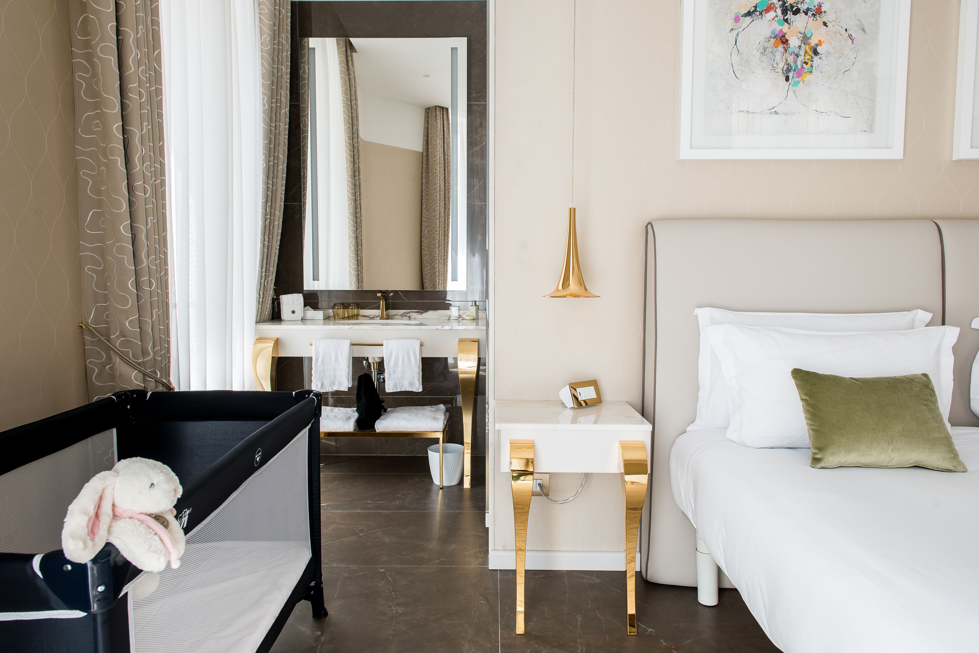 A hotel room with a crib, a bedside table, a bed, a mirror, and a bathroom with a sink and towel rack.