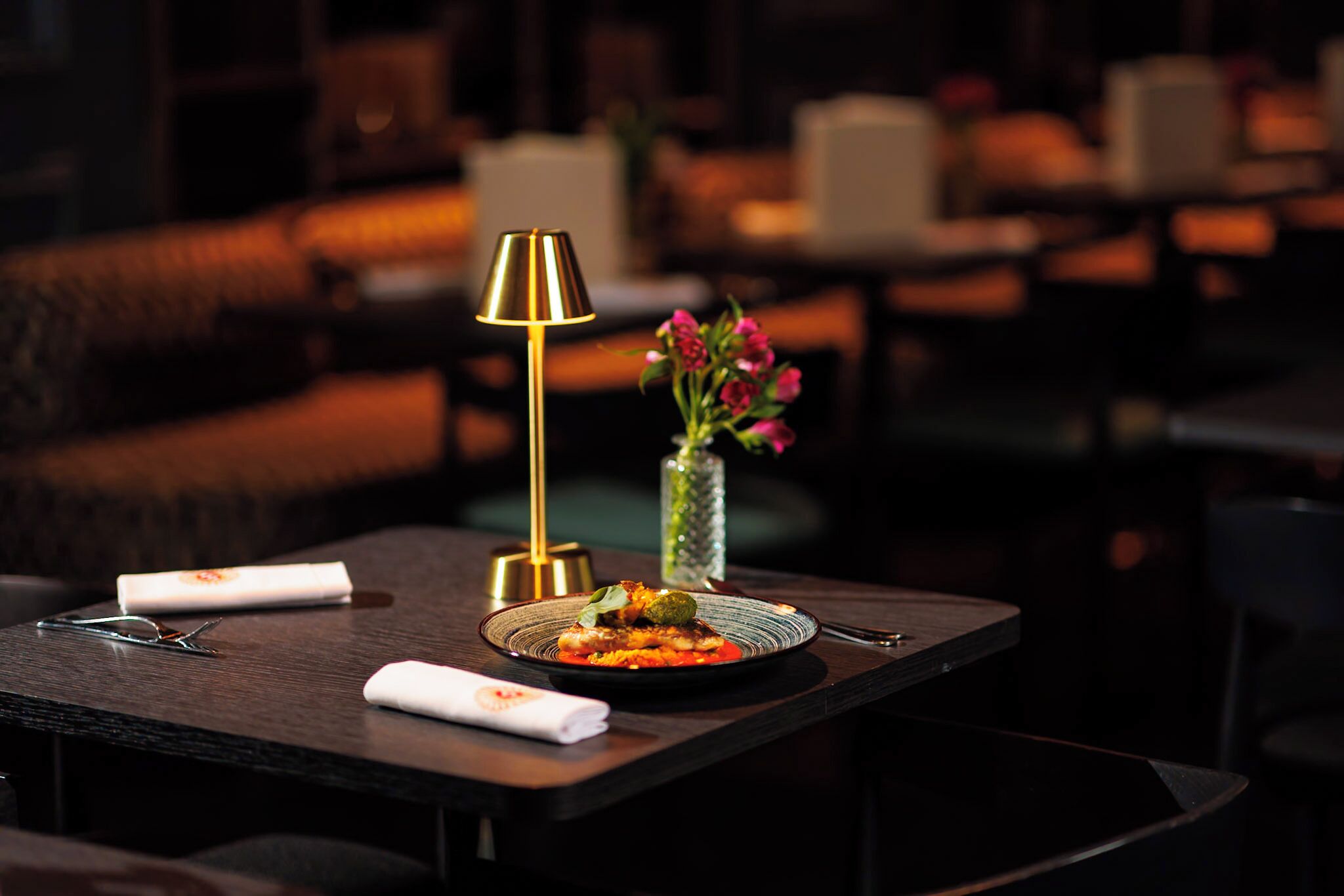 Une table à manger avec une assiette de nourriture, un vase rempli de fleurs et une lampe.