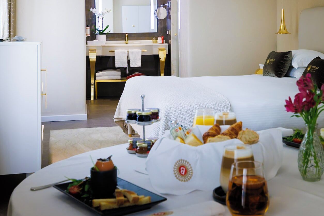 Une chambre d’hôtel comprenant un lit, une table à manger avec des aliments pour le petit-déjeuner, une salle de bain avec un miroir et un vase avec des fleurs.