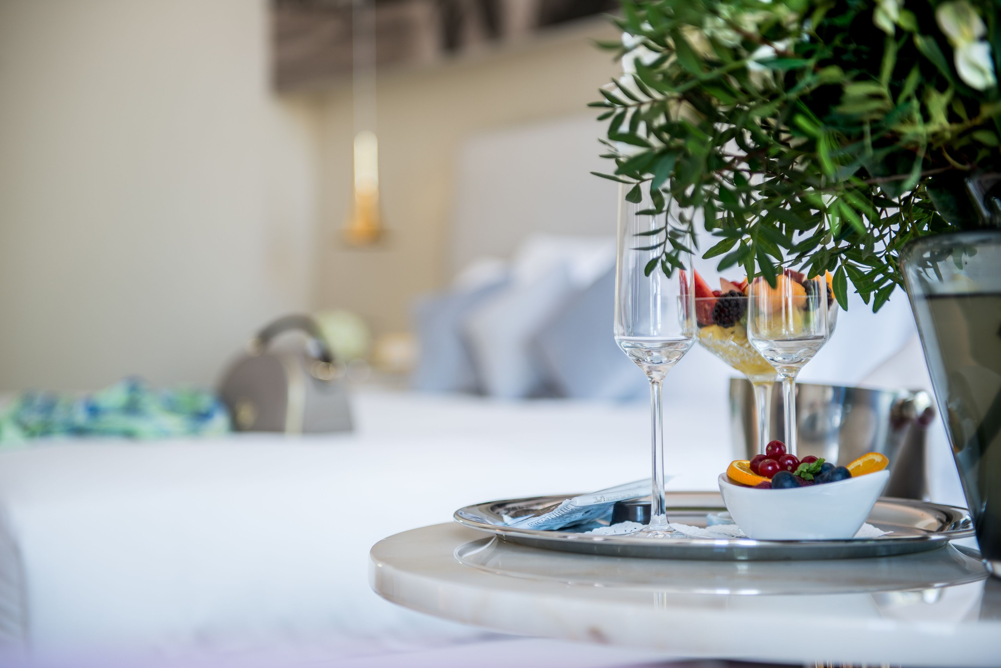 Un plateau avec un saladier de fruits et deux verres contenant un liquide clair, peut-être du champagne, est posé sur une table ronde recouverte d’une nappe blanche.