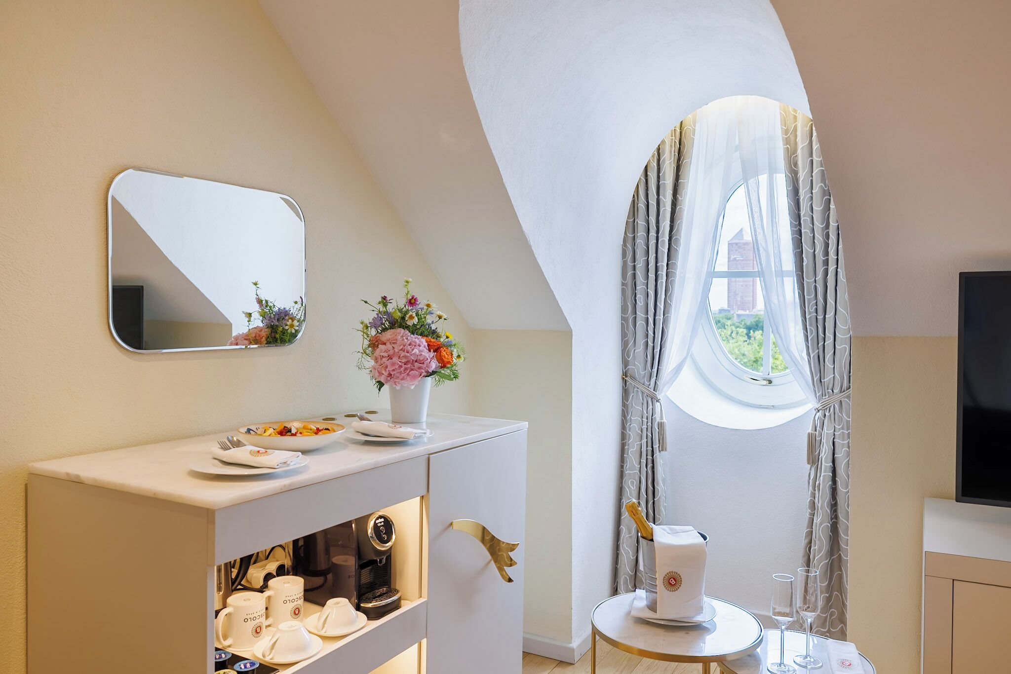 A modern bathroom with a vanity, a mirror, a window with curtains, a coffee machine, and a small table with a glass and a bottle.