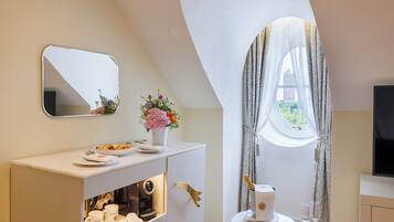 A modern bathroom with a vanity, a mirror, a window with curtains, a coffee machine, and a small table with a glass and a bottle.