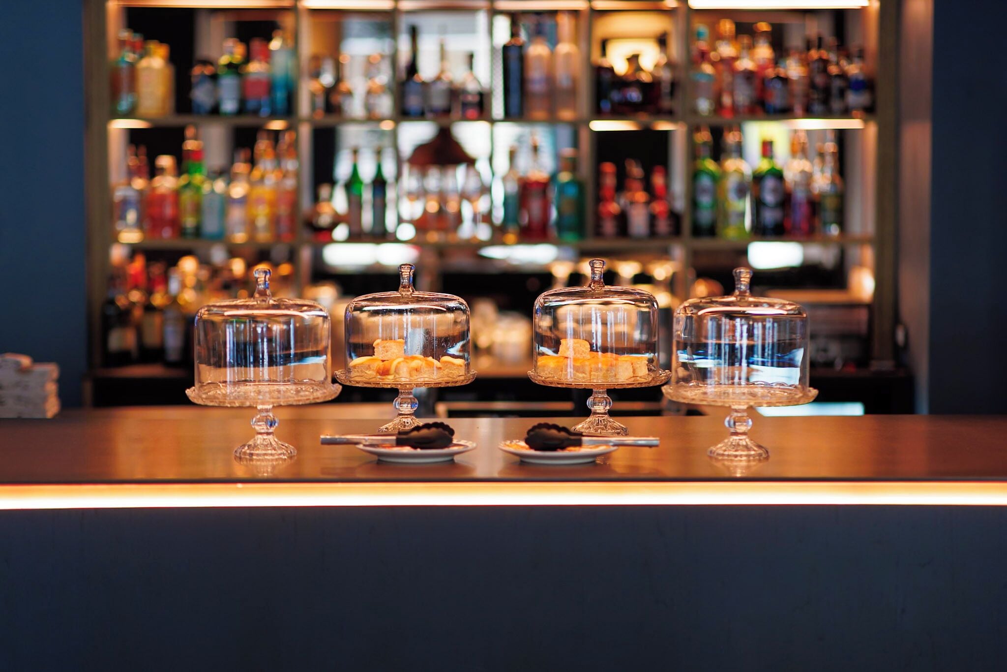 A bar counter with three glass-covered dessert displays and a background of shelves stocked with various bottles.