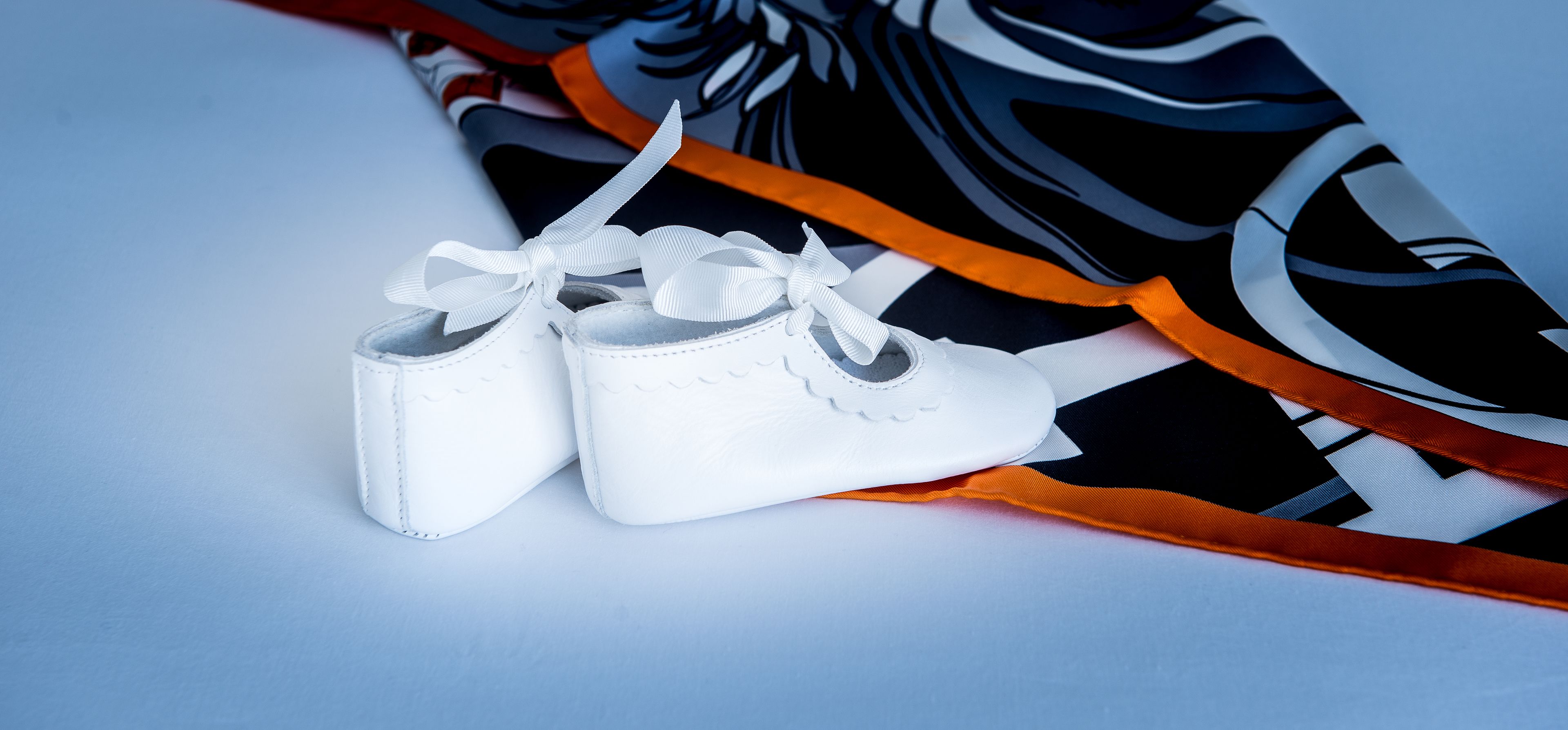 A pair of white baby shoes with a bow, placed on a surface with a black and white patterned fabric and an orange ribbon.