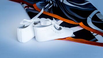 A pair of white baby shoes with a bow, placed on a surface with a black and white patterned fabric and an orange ribbon.