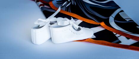 A pair of white baby shoes with a bow, placed on a surface with a black and white patterned fabric and an orange ribbon.