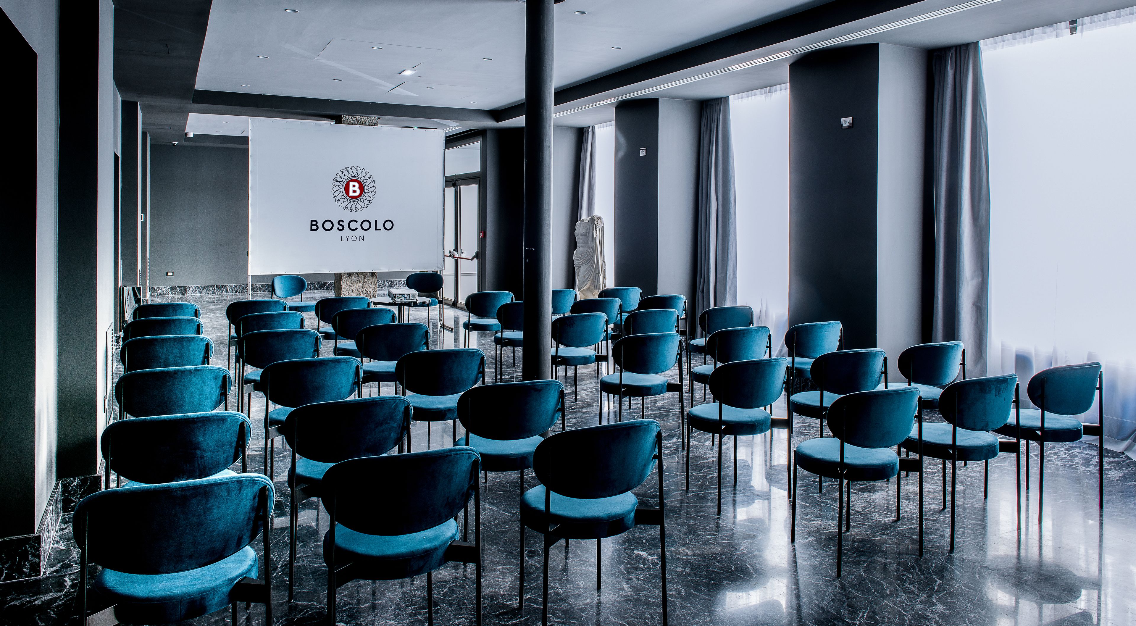 Un salón de conferencias con filas de sillas azules, un gran cartel blanco con el logotipo y el nombre 'BOSCOLO LYC' en la pared, y una estatua al fondo.