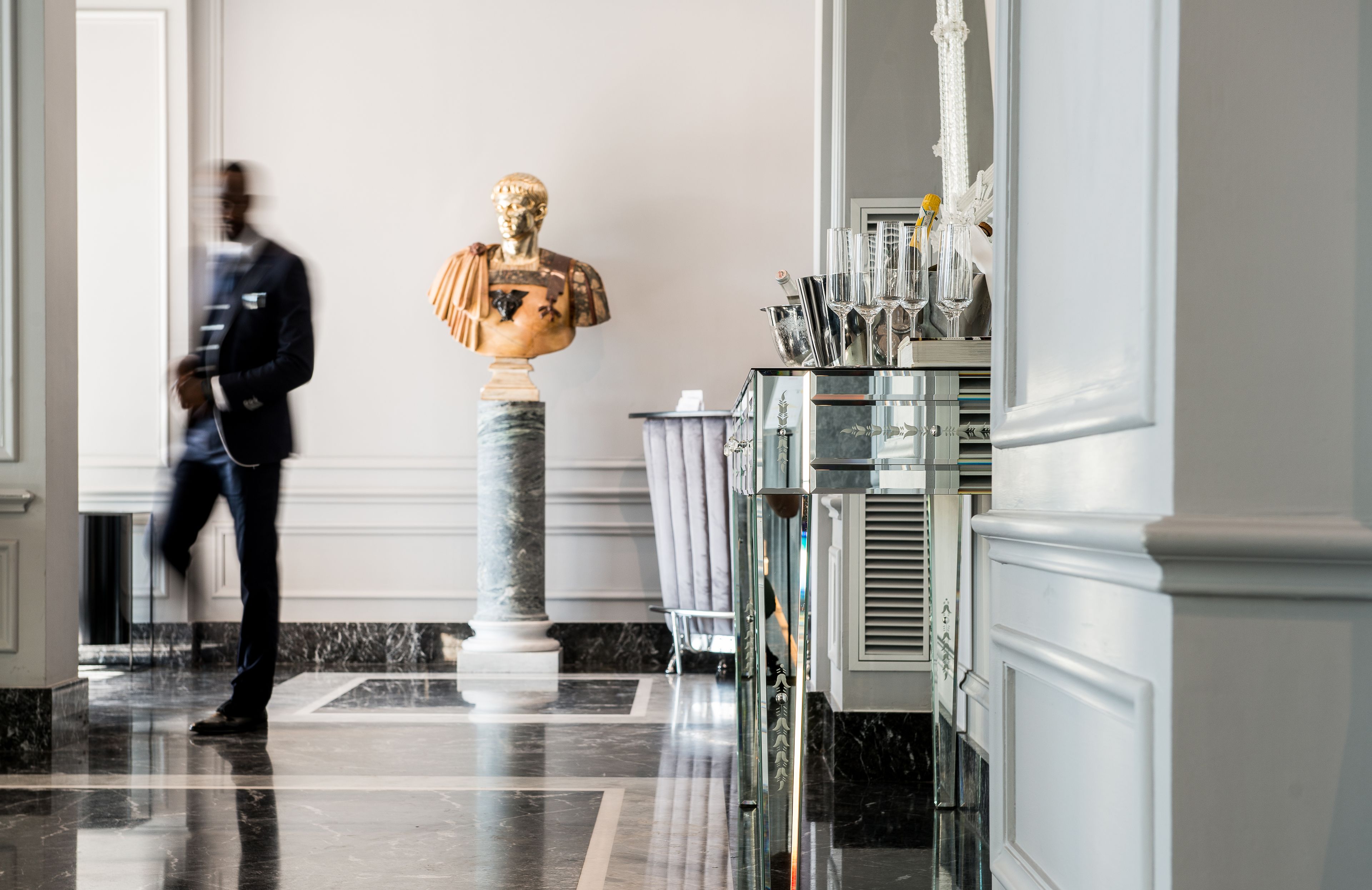 A marble lobby with a bust sculpture, a glass cabinet with silverware, and a person walking.