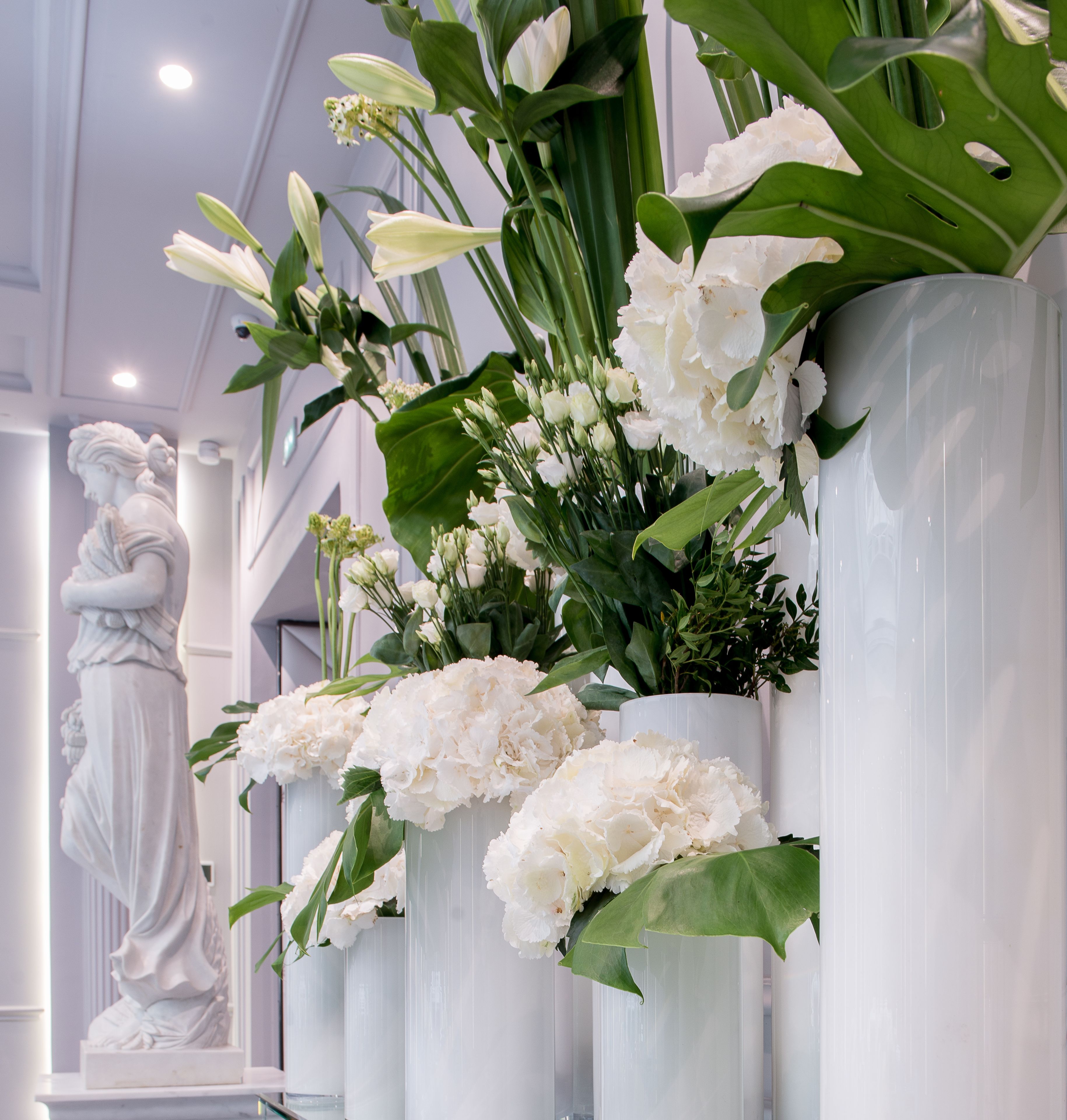 A statue of a woman in a classical pose, surrounded by white flowers in white vases.