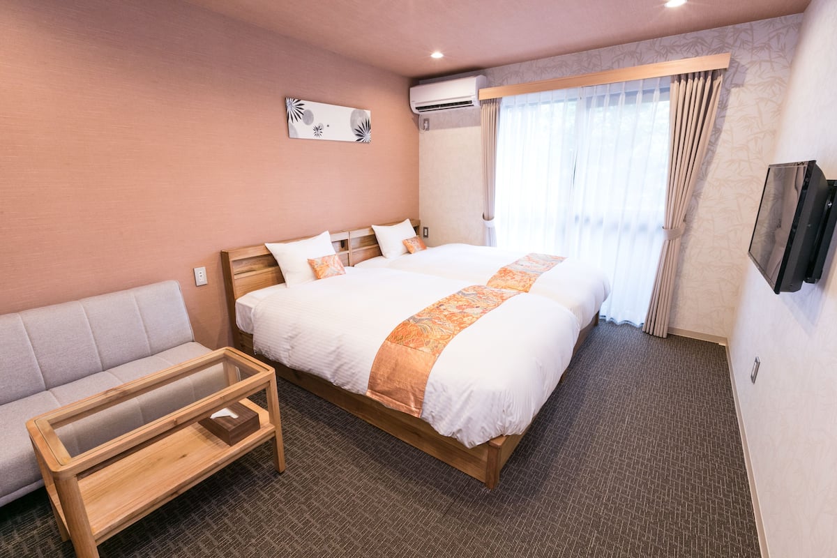 A hotel room with a large bed, a sofa, a wooden coffee table, an air conditioning unit, and a TV mounted on the wall.