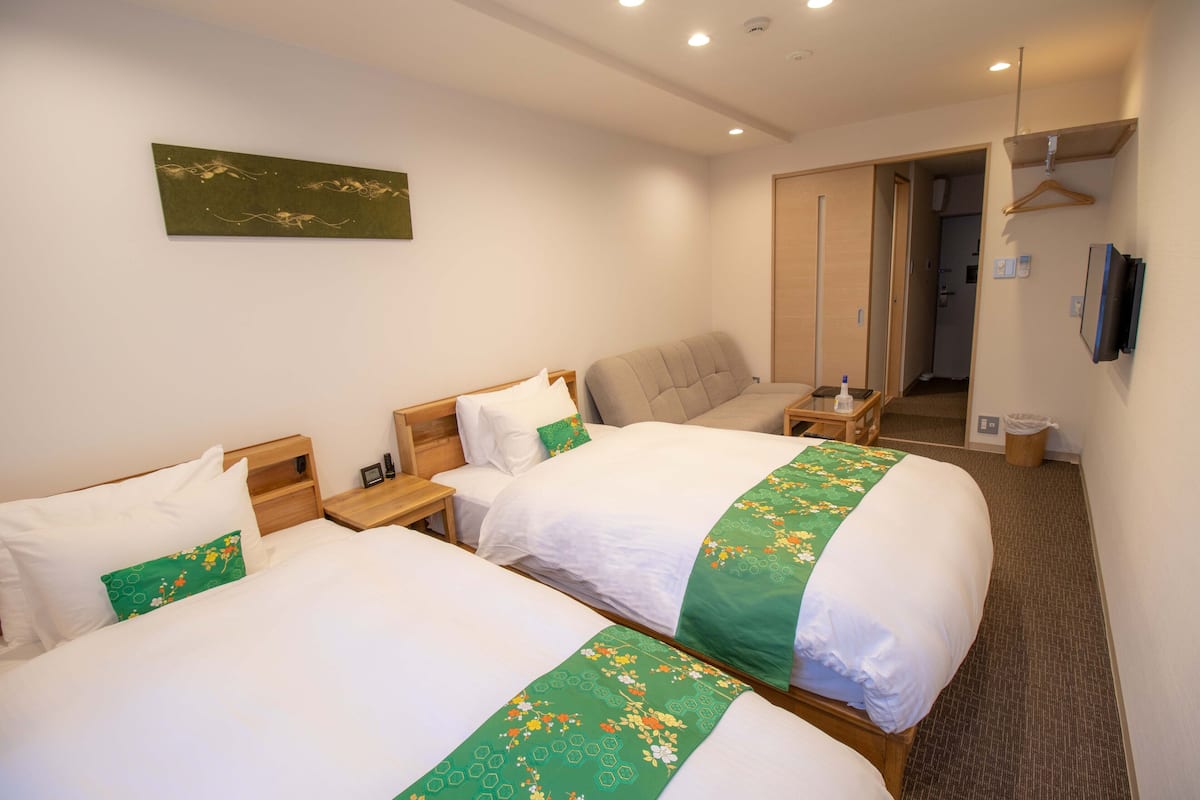 A hotel room with two beds, a wooden headboard, a bedside table, a green floral bedspread, a wall-mounted TV, and a door leading to a hallway.