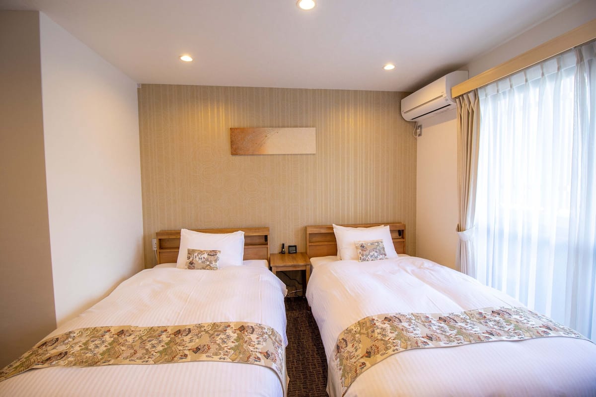 A hotel room with two beds, a nightstand, an air conditioning unit, and a window with curtains.