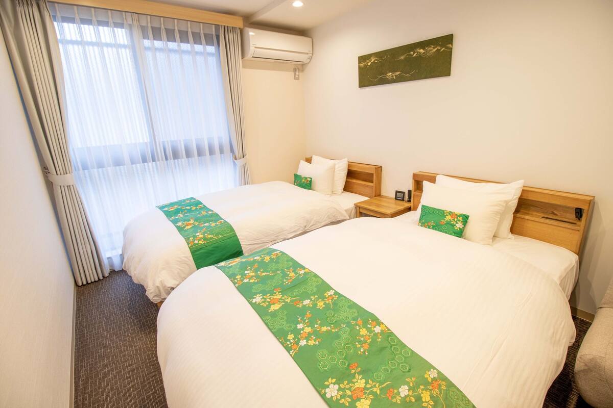 A hotel room with two beds, a window with curtains, an air conditioner, and a wall-mounted artwork.