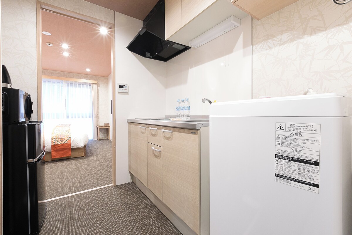 Cocina pequeña con refrigerador, pileta y microondas. Hay una ventana con cortinas y una cama en la habitación contigua.