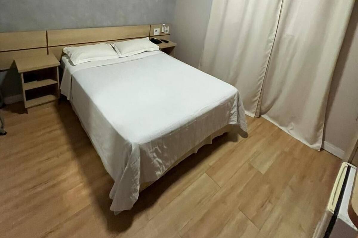 A bedroom with a bed, a nightstand, a desk, an air conditioning unit, and a curtain.