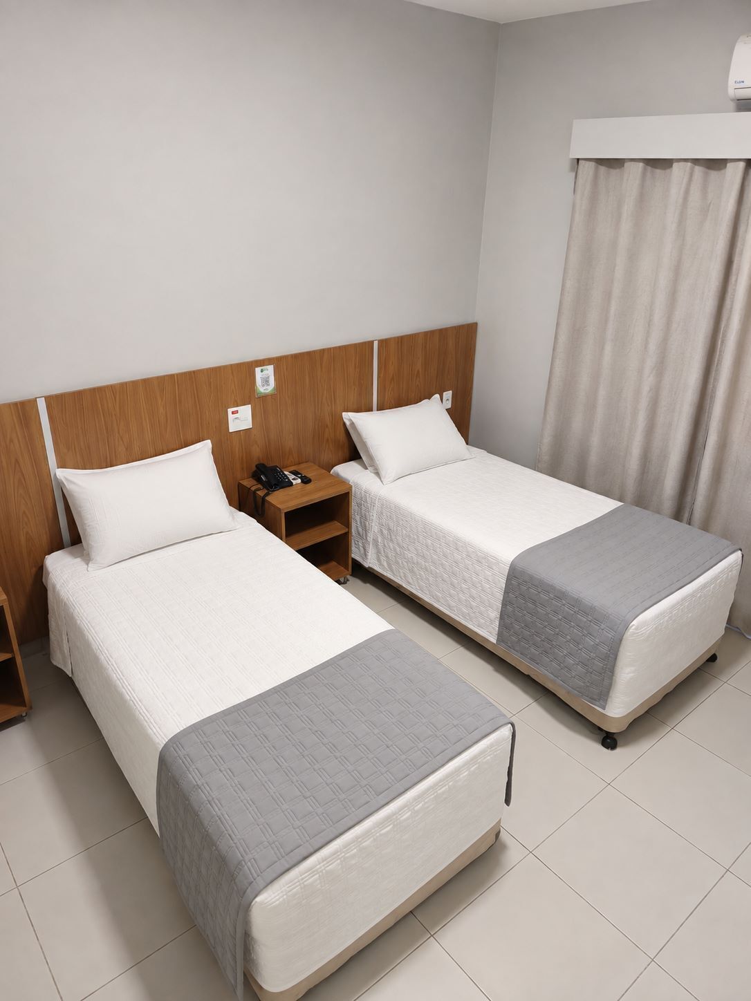 A hotel room with two single beds, a wooden headboard, a nightstand with a phone, and a window with curtains.