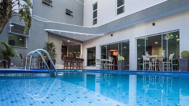 An indoor swimming pool with clear blue water, surrounded by a modern building with large windows and a seating area with chairs and tables. - Hotel Serra de Jundiaí (Jundiai)