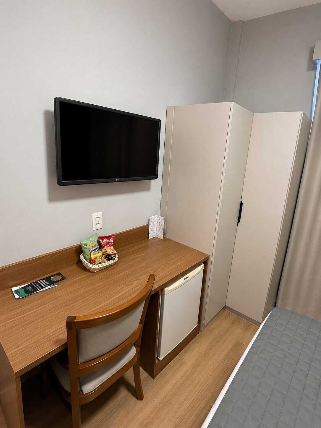 A compact hotel room with a wooden desk, a television mounted on the wall, a small refrigerator, and a bed with a grey quilt.