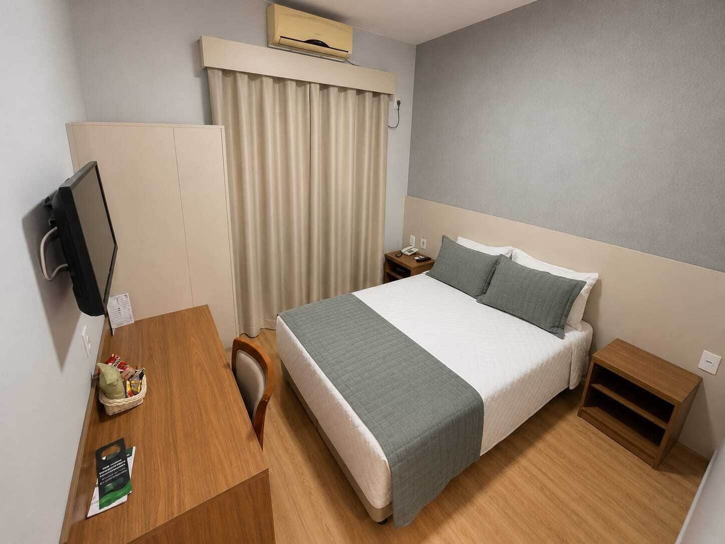 Quarto de hotel com cama, mesas de cabeceira, escrivaninha com cadeira, televisão e ar condicionado.