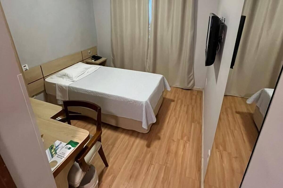 A compact hotel room with a single bed, a small table, a chair, a bedside table, a TV, and an air conditioning unit.