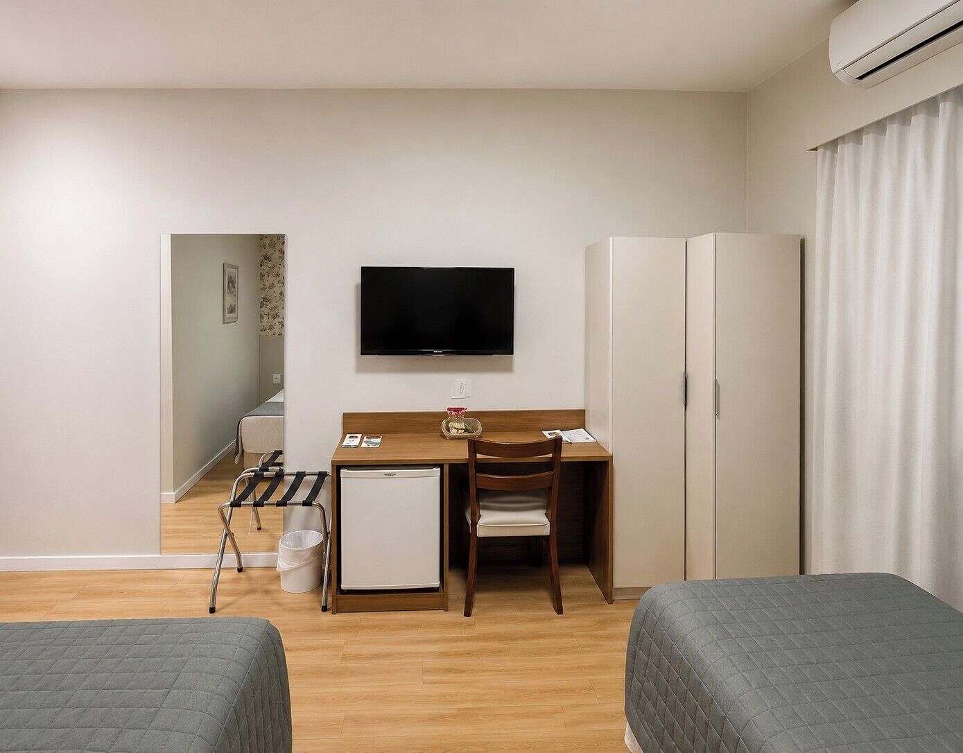 Quarto de hotel com uma cama, uma escrivaninha, uma cadeira, uma televisão, um guarda-roupa e um pequeno frigorífico.
