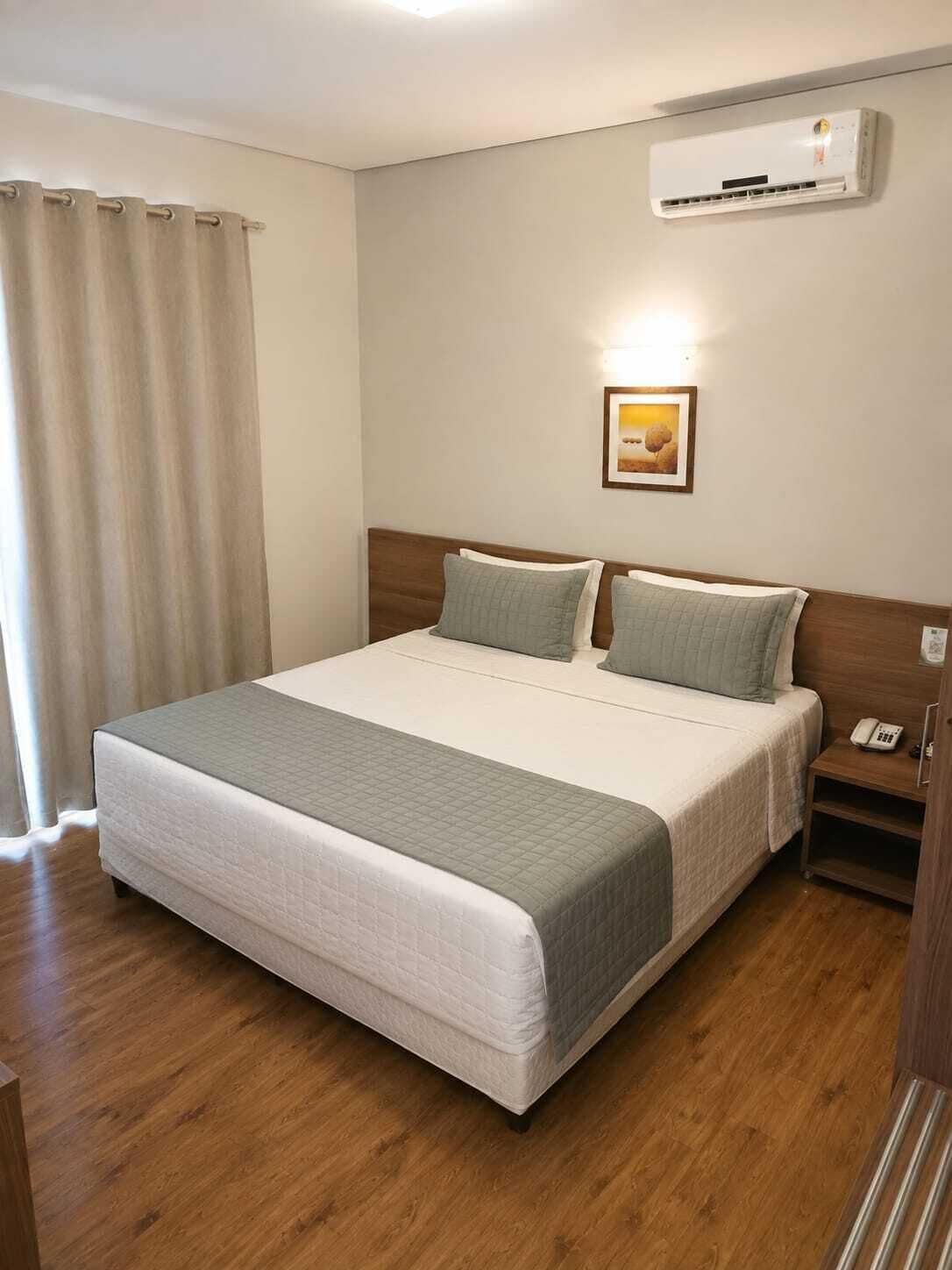 Habitación de hotel con cama, mesita de noche, teléfono, unidad de aire acondicionado y ventana con cortinas.