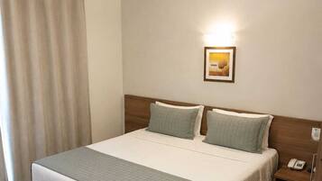 Habitación de hotel con cama, mesita de noche, teléfono, unidad de aire acondicionado y ventana con cortinas.