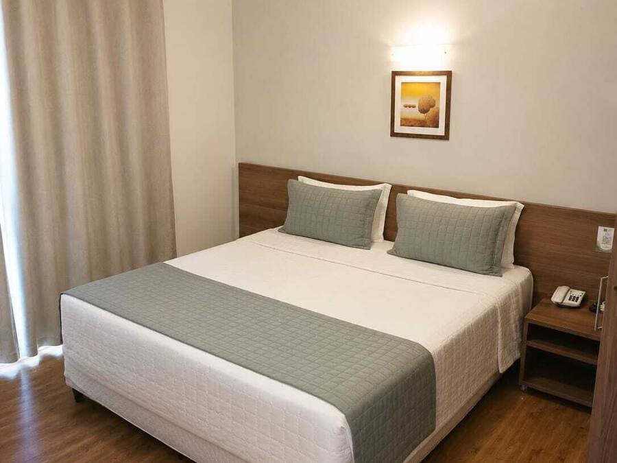Habitación de hotel con cama, mesita de noche, teléfono, unidad de aire acondicionado y ventana con cortinas.