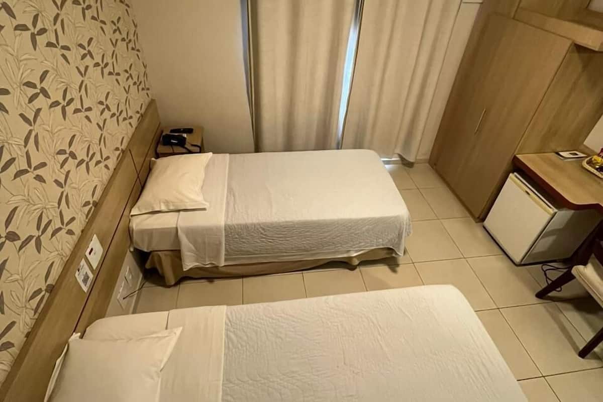 A hotel room with two single beds, a wooden wardrobe, a desk with a chair, and an air conditioning unit.