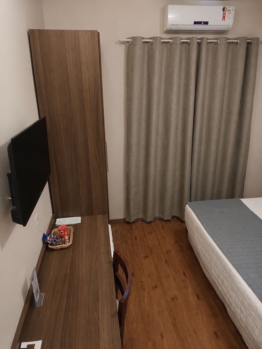 Une pièce comprenant un bureau en bois, un téléviseur à écran plat, un lit avec un matelas gris et une fenêtre ornée de rideaux beige.