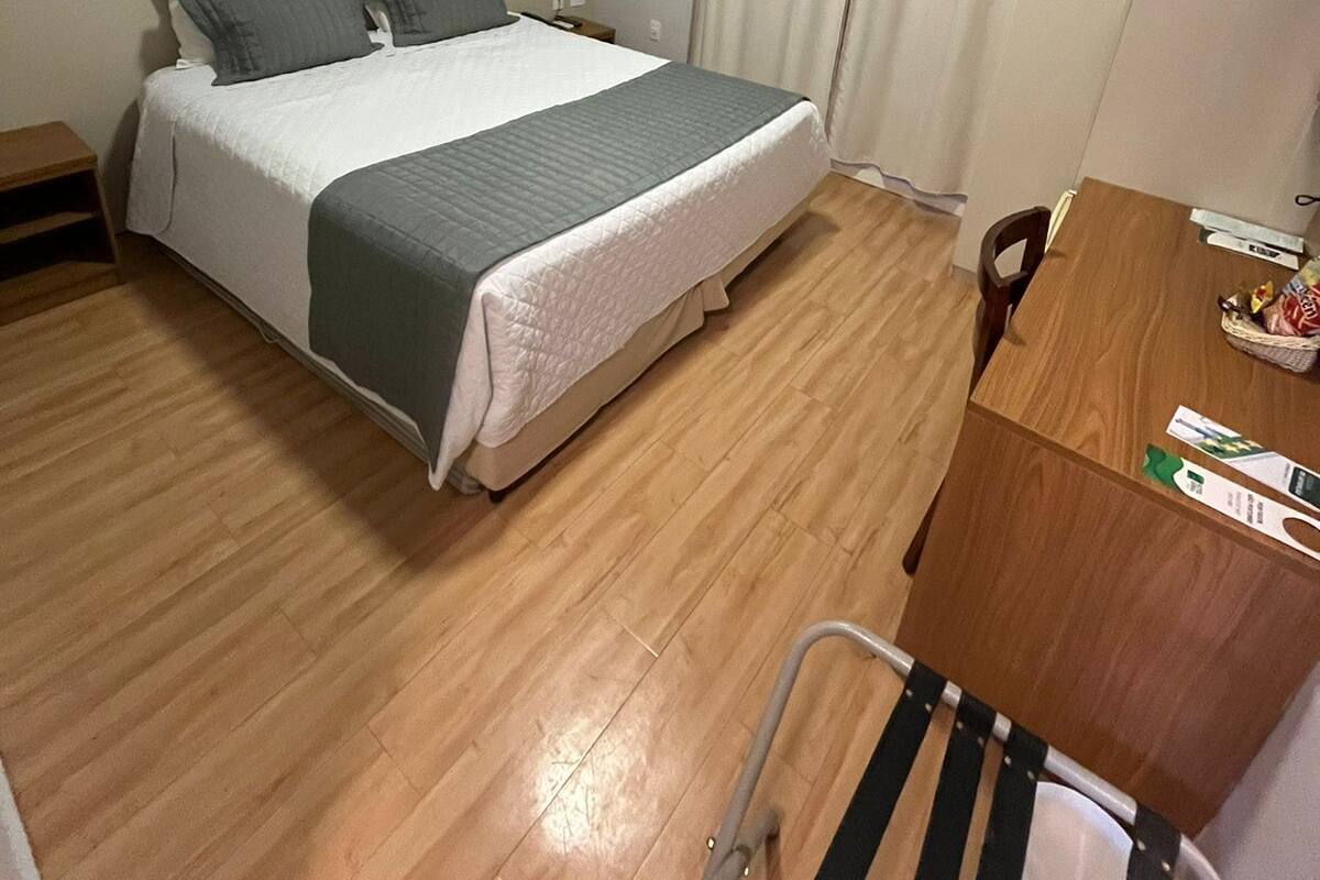 A hotel room with a bed, a desk, and a chair. There is an air conditioning unit above the bed and a window with curtains.