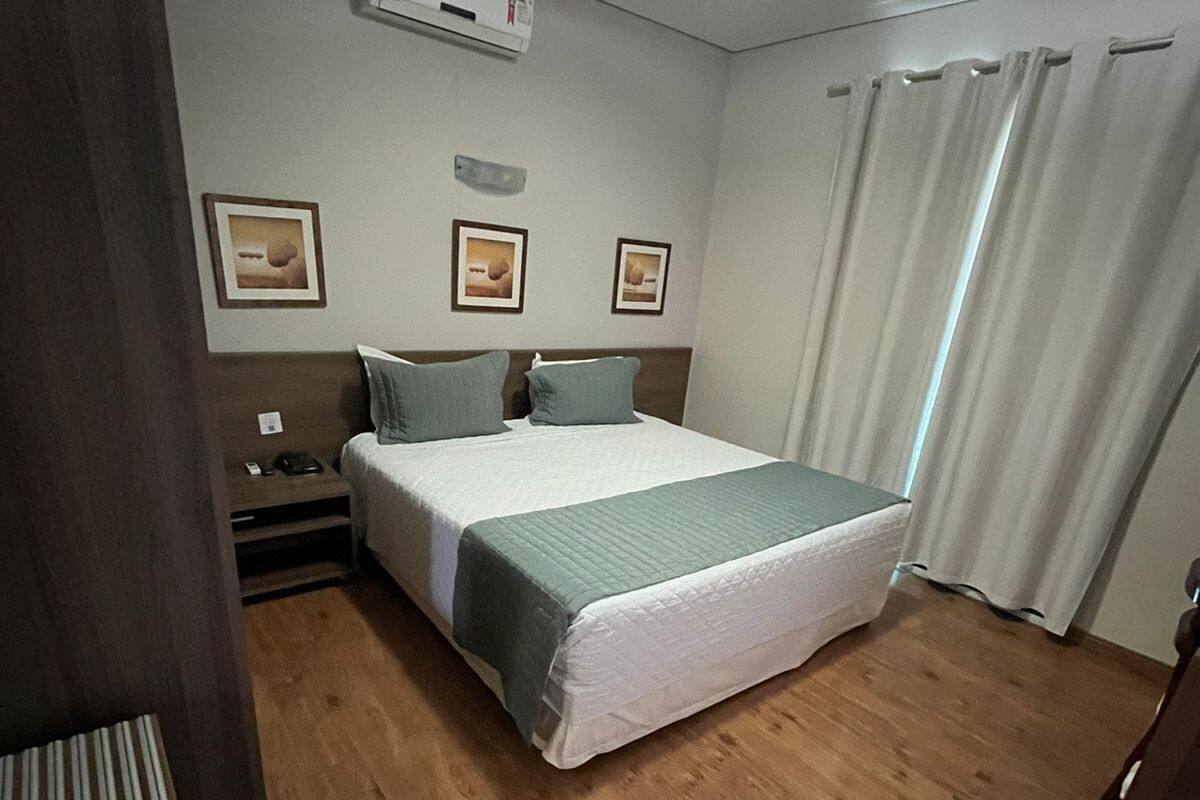 A hotel room with a bed, a nightstand, an air conditioning unit, and a window with curtains.