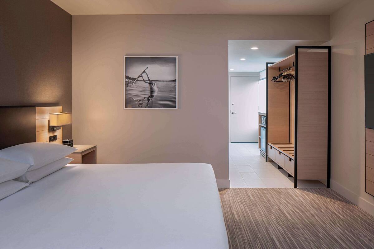 A hotel room with a bed, a nightstand with a lamp, a wooden wardrobe, and a framed picture on the wall.