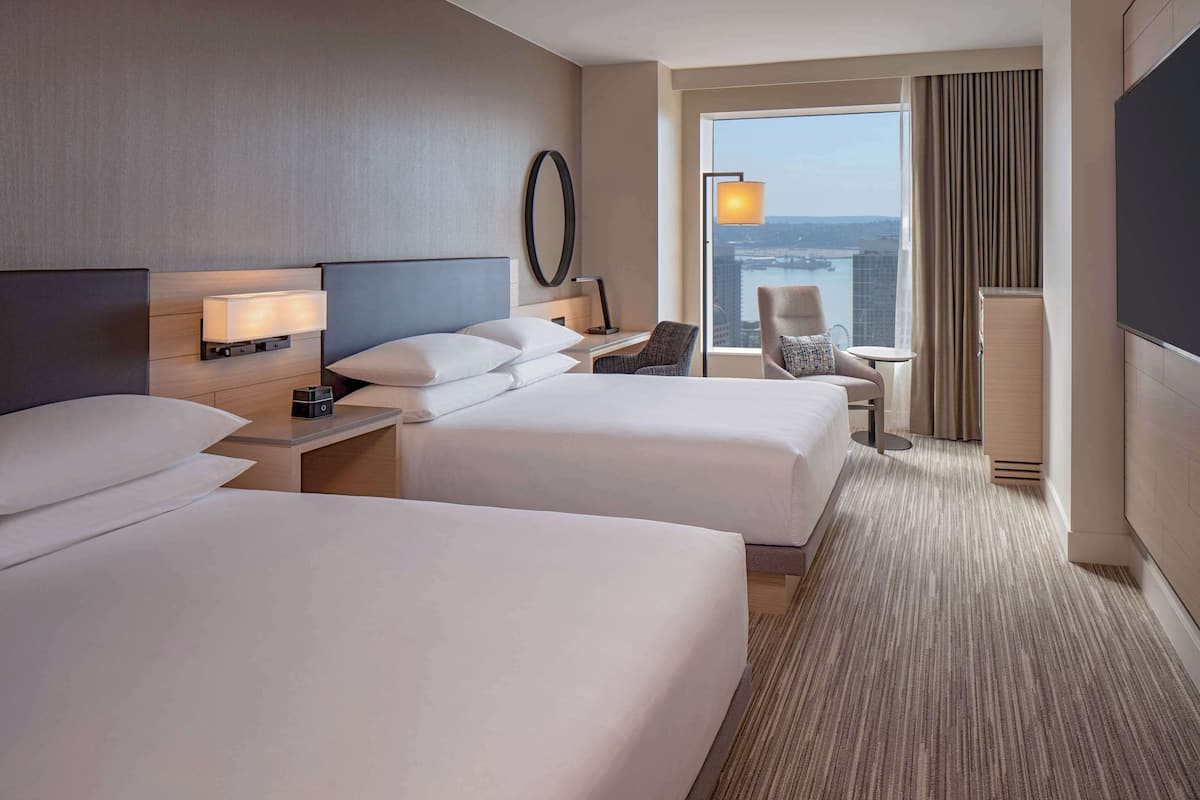 A hotel room with two beds, a desk, a chair, a TV, and a large window with a city view.