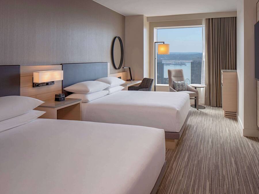 Habitación de hotel con dos camas, un escritorio, una silla, un televisor y un ventanal con vistas a la ciudad.