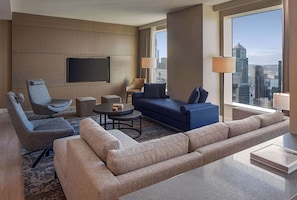 A modern hotel room with a large window offering a city view, a flat-screen TV mounted on the wall, a grey armchair, a blue sofa, and a beige sofa.